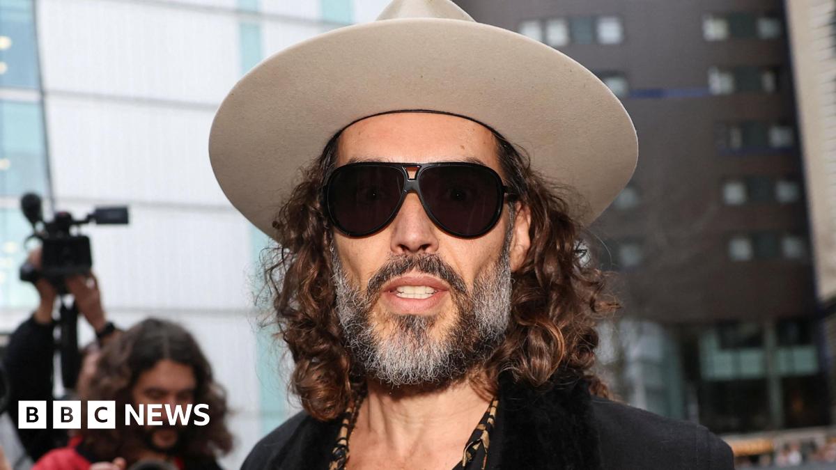 Russell Brand arriving at Southwark Crown Court wearing a light grey fedora hat, dark glasses, a leopard print shirt unbuttoned halfway down his chest, a cross on a necklace, and a dark jacket