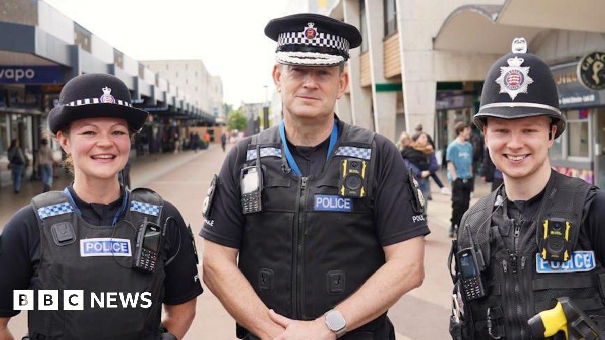 Chief Constable Ben-Julian Harrington with fellow police officers either side of him. All are wearing black police uniform and hats while standing in Basildon town centre, which is lined with shops.