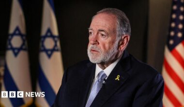 Mike Huckabee, a man with short grey hair and facial hair, sits facing the left of  the frame in a dark room. Behind him are two Israeli flags and one US flag. He wears a black suit jacket, a white shirt, blue tie, and a yellow ribbon pin.