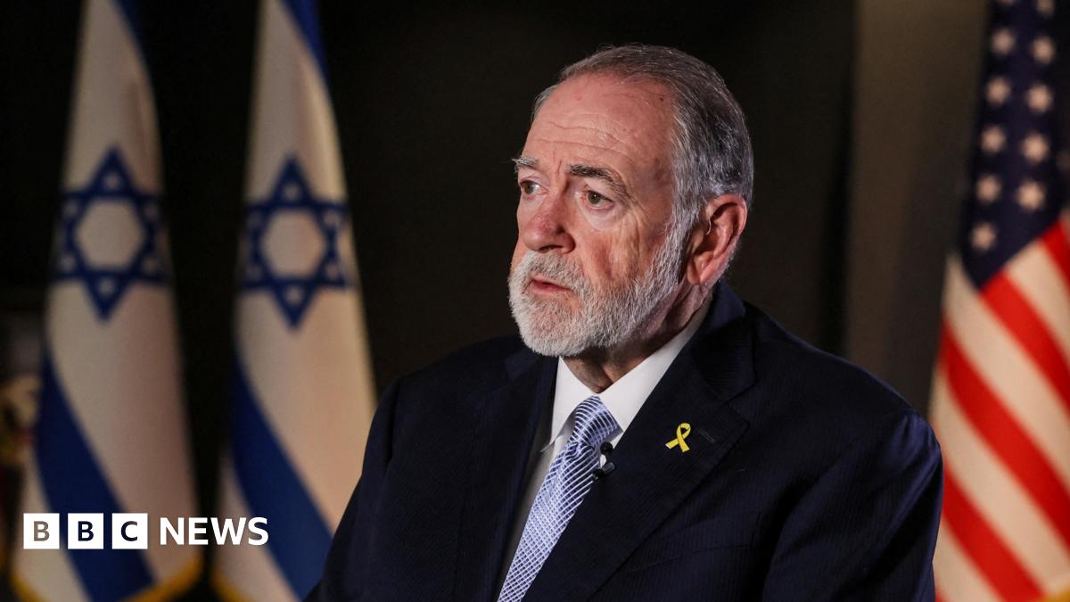 Mike Huckabee, a man with short grey hair and facial hair, sits facing the left of  the frame in a dark room. Behind him are two Israeli flags and one US flag. He wears a black suit jacket, a white shirt, blue tie, and a yellow ribbon pin.
