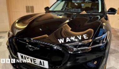 A Wave car sits in a showroom, the numberplate reads "Wayve dot AI". The car is black and has white lettering on the bonnet which says "WAYVE".