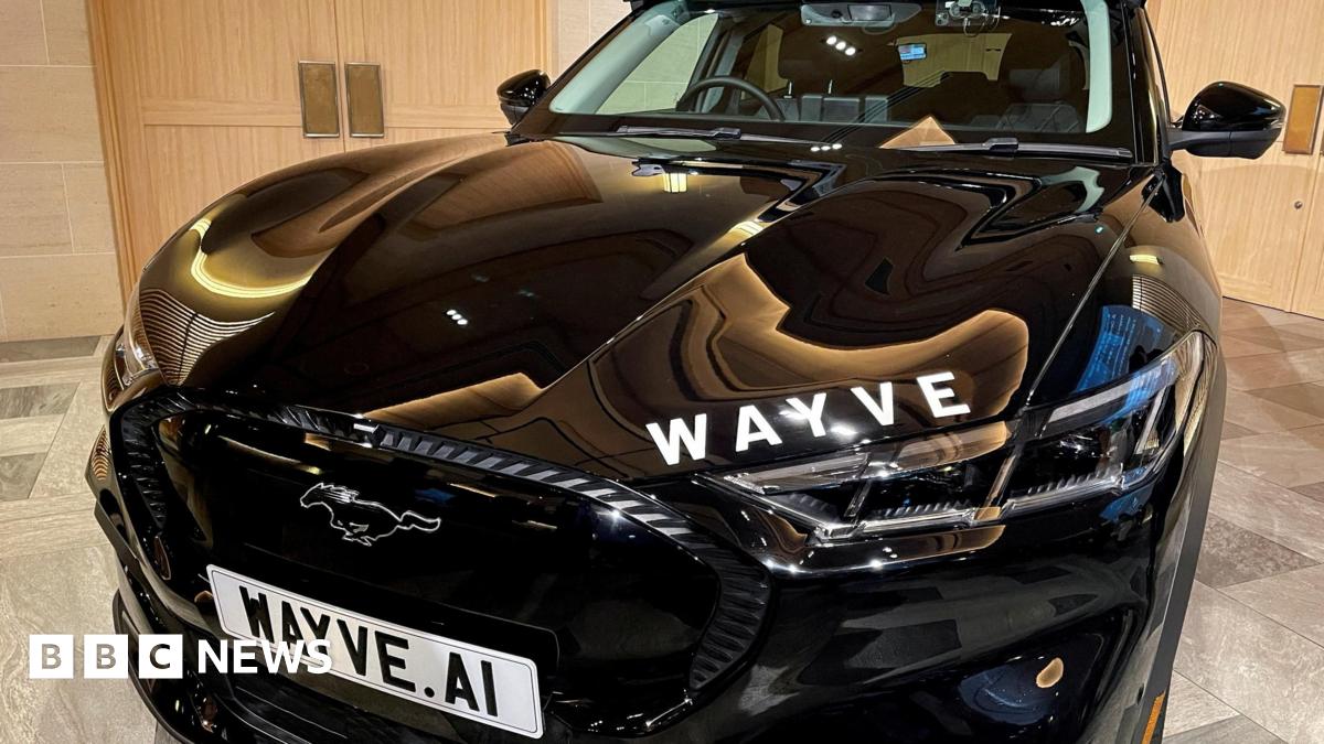 A Wave car sits in a showroom, the numberplate reads "Wayve dot AI". The car is black and has white lettering on the bonnet which says "WAYVE".