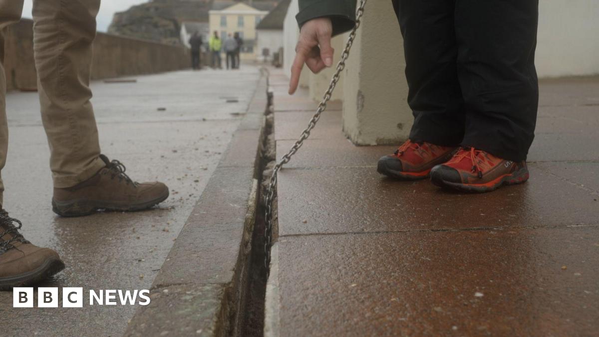 Two people are standing on either side of the long crack in the ground. A person wearing brown hiking boots is stood on the left side of the crack, while a person wearing red walking boots is standing on the right side of the crack. A metal chain leads into the crack. A hand is pointing at the crack.