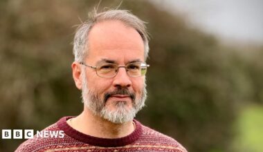 A man stands outside with a large hedge blurred in the background. He is wearing wire rimmed glasses, has short grey hair and a beard. He is wearing a patterned red jumper.
