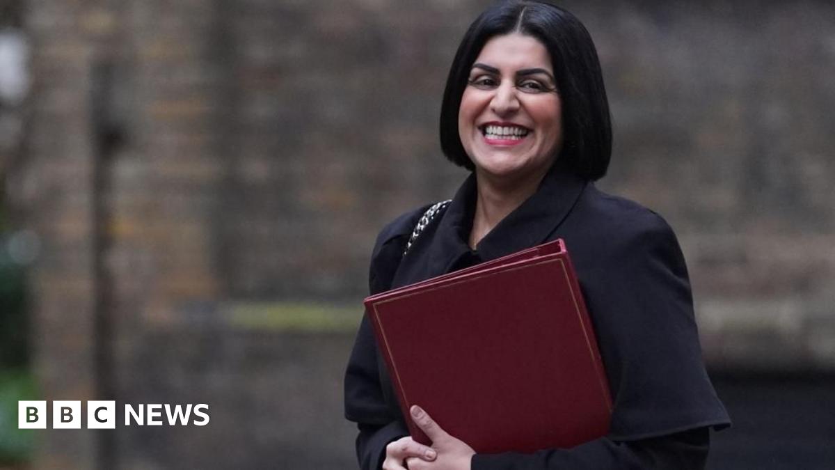 Shabana Mahmood is smiling as she walks in Downing Street. She is holding a red folder and wearing a dark coloured coat.