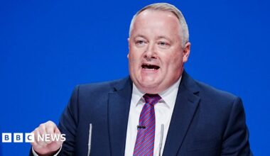 Darren Millar is wearing a blue suit, striped red and blue tie and a white shirt, and is making a speech on stage in front of a blue background