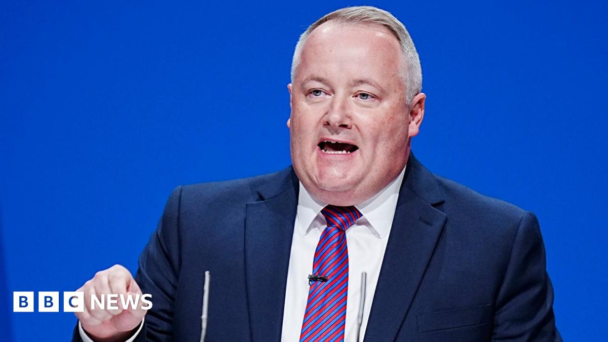 Darren Millar is wearing a blue suit, striped red and blue tie and a white shirt, and is making a speech on stage in front of a blue background