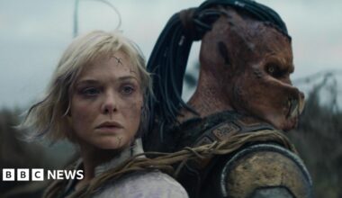 A human-like figure with blond hair and blue scars on her face appears strapped back to back with a humanoid alien-like creature, with orange skin and  insect-like jaws over its mouth.