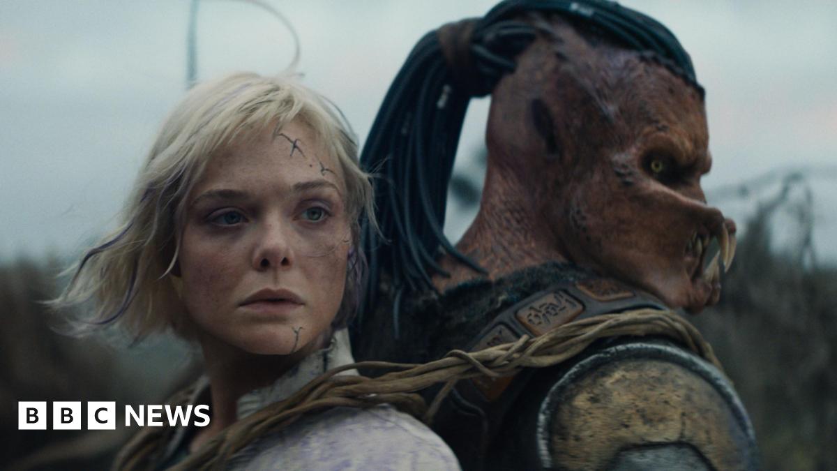 A human-like figure with blond hair and blue scars on her face appears strapped back to back with a humanoid alien-like creature, with orange skin and  insect-like jaws over its mouth.