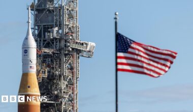 Orange and white Artemis rocket next to a towering grey space system and a blue, white, and red starts and stripes flag, against a blue sky