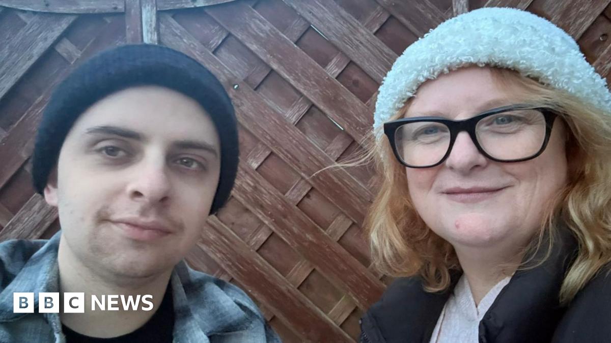 A young man and a middle-aged woman - Noah Herniman and his mother Shelley - stand next to each other next to a garden fence, both wearing beanie hats.