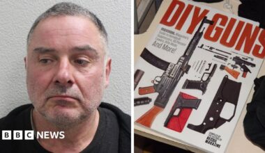 Thomas McKenna is smiling and is holding a replica AK47 assault rifle. He is wearing a black hoodie and blue jeans. There are bushes on either side. There are two people you can barely see behind him in brightly coloured clothing.