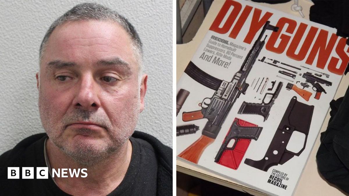 Thomas McKenna is smiling and is holding a replica AK47 assault rifle. He is wearing a black hoodie and blue jeans. There are bushes on either side. There are two people you can barely see behind him in brightly coloured clothing.