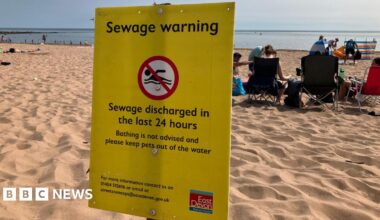 A beach scene with several people sitting on the sand facing the sea. In the foreground, a prominent yellow warning sign from East Devon District Council advises against bathing due to recent sewage discharge and warns to keep pets out of the water.