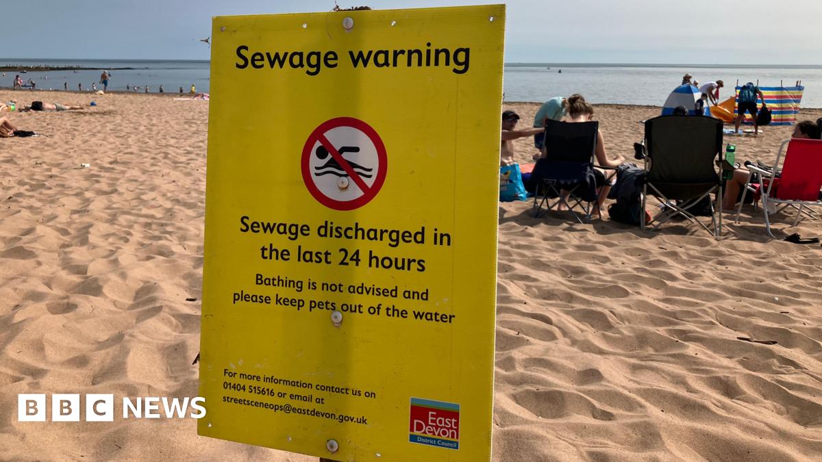 A beach scene with several people sitting on the sand facing the sea. In the foreground, a prominent yellow warning sign from East Devon District Council advises against bathing due to recent sewage discharge and warns to keep pets out of the water.
