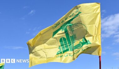 The yellow and green Hezbollah flag flies in the breeze, against a clear blue sky.