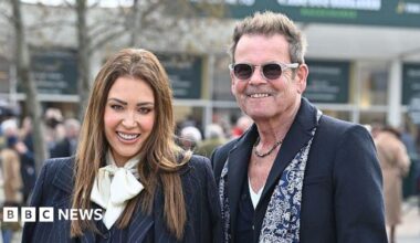A woman with long-brown hair and wearing a dark pinstriped suit with a white scarf stands next to a man with a dark coloured suit and blue and white patterned scarf. He has short grey hair which is swept back and spiked up. He has a chain necklace and sunglasses. They are both smiling.