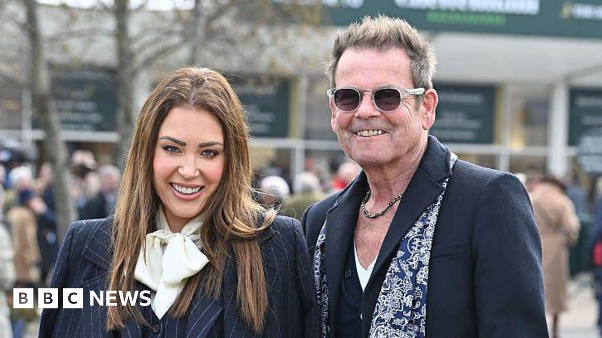 A woman with long-brown hair and wearing a dark pinstriped suit with a white scarf stands next to a man with a dark coloured suit and blue and white patterned scarf. He has short grey hair which is swept back and spiked up. He has a chain necklace and sunglasses. They are both smiling.