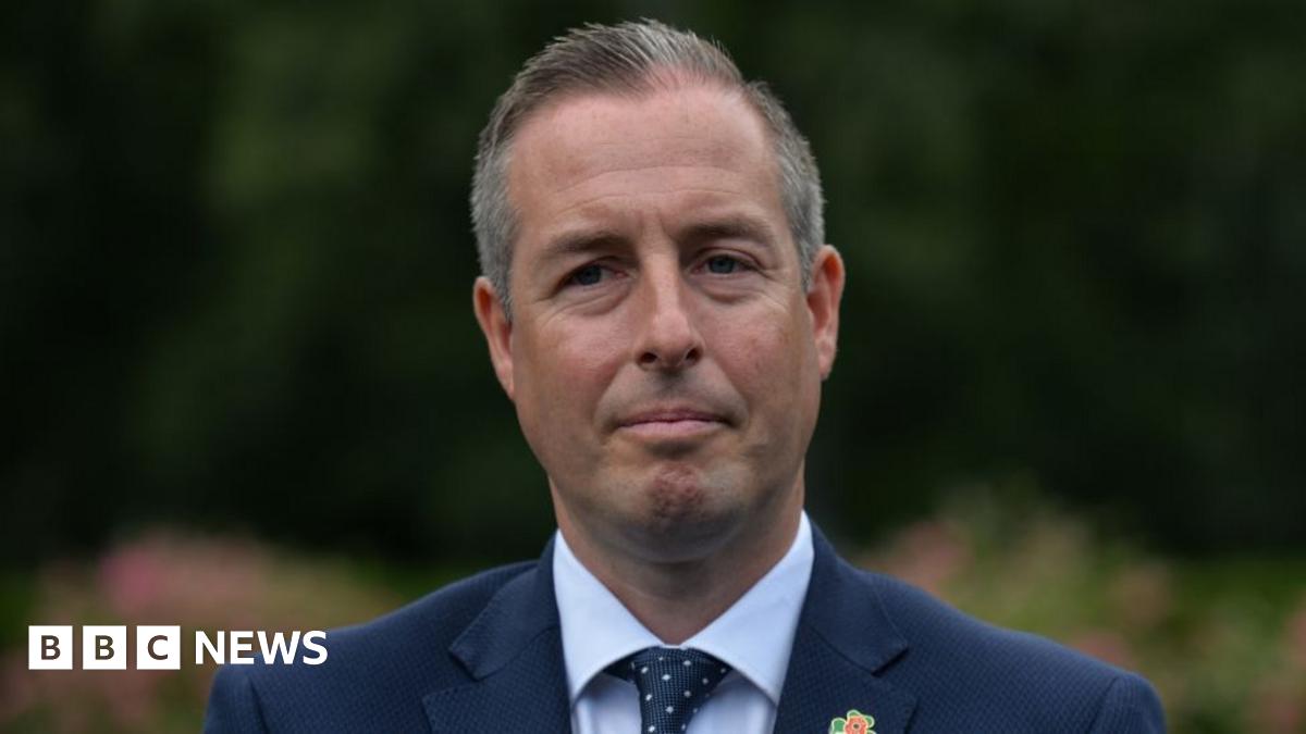 Paul Givan stood outside with a neutral expression.   He has short, greying hair and is wearing a navy suit, a white shirt and a navy spotted tie.