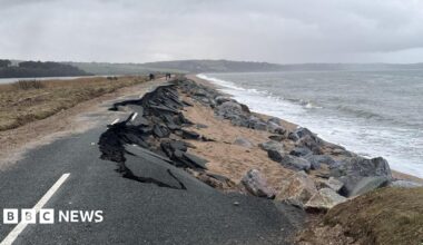 The A379 Slapton Line between Torcross and Slapton. The road is damaged with one lane washed away. The sea is to the right and there is a grassy patch and a lake to the left.