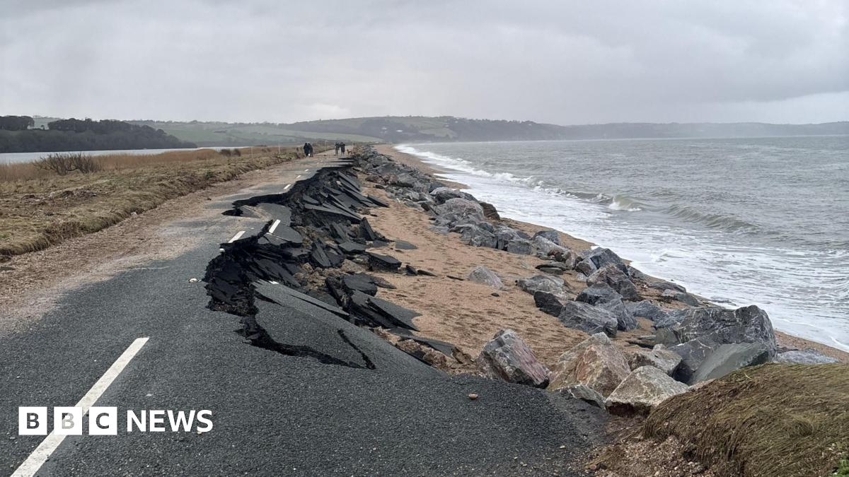 The A379 Slapton Line between Torcross and Slapton. The road is damaged with one lane washed away. The sea is to the right and there is a grassy patch and a lake to the left.