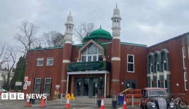 The entrance to a mosque, a brick building with dark green signage. The is a green dome shaped structure on the roof and two minaret towers with Islamic crescent moon sculptures on top.