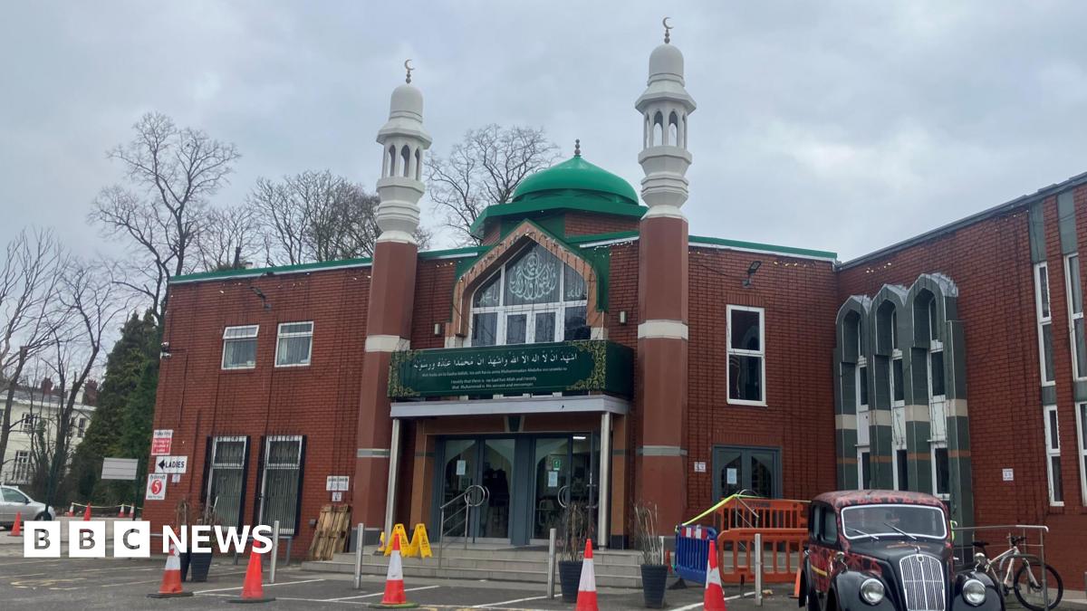 The entrance to a mosque, a brick building with dark green signage. The is a green dome shaped structure on the roof and two minaret towers with Islamic crescent moon sculptures on top.