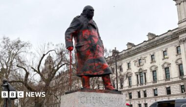 Winston Churchill statue seen from the left front side, looking up at the statue,  covered in red paint. Buildings in the background