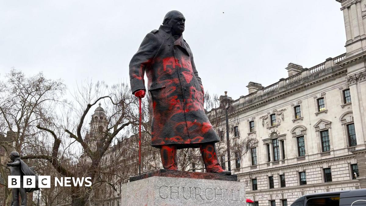 Winston Churchill statue seen from the left front side, looking up at the statue,  covered in red paint. Buildings in the background