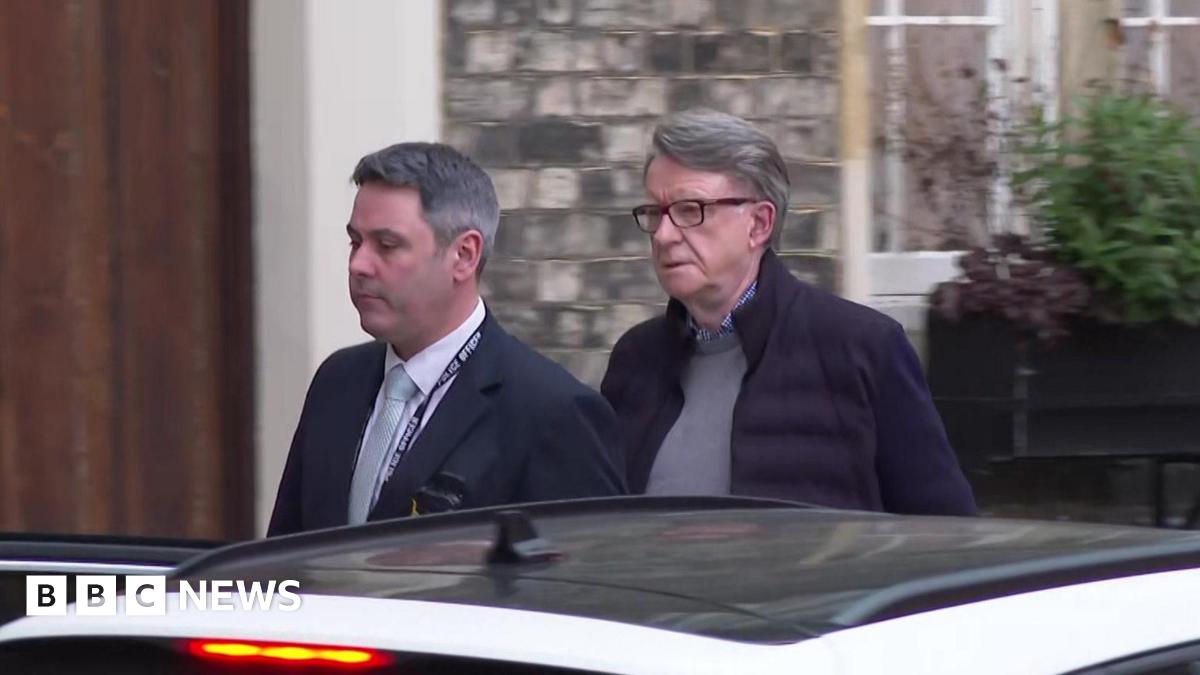 Lord Mandelson leaves his London home, walking behind a police officer wearing a suit, towards a white car with the door open.