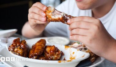 Person in a white t-shirt eating sticky chicken wings with their hands from a white bowl on top of a larger plate where they have left the bones. The person's hands have a bit of sauce on them and their face is not visible.