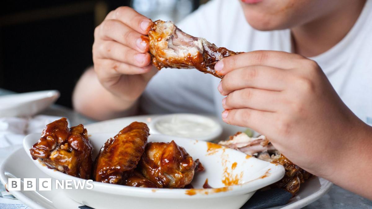 Person in a white t-shirt eating sticky chicken wings with their hands from a white bowl on top of a larger plate where they have left the bones. The person's hands have a bit of sauce on them and their face is not visible.