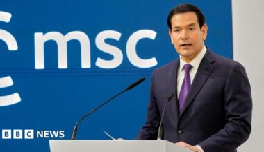 Marco Rubio stands at a podium with two microphones, addressing an audience against a blue backdrop displaying the letters “msc”.