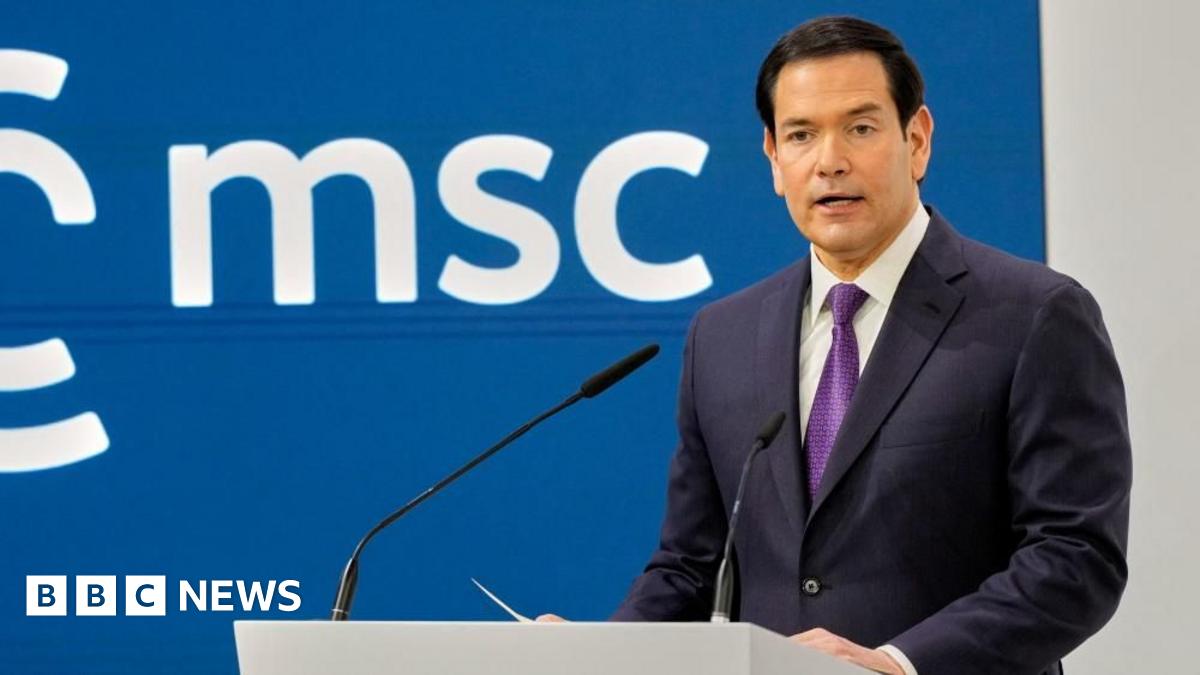 Marco Rubio stands at a podium with two microphones, addressing an audience against a blue backdrop displaying the letters “msc”.