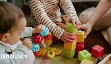 High angle shot of children's hands holding multi-coloured bricks and rattle while adult hand assisting them to build tower