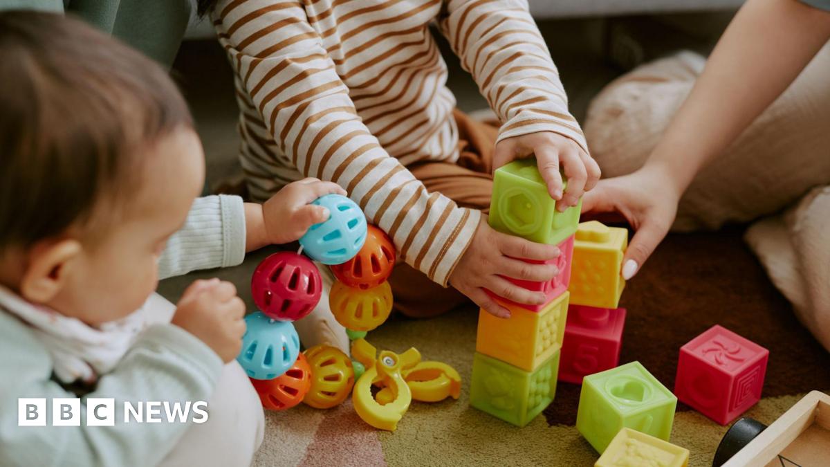 High angle shot of children's hands holding multi-coloured bricks and rattle while adult hand assisting them to build tower