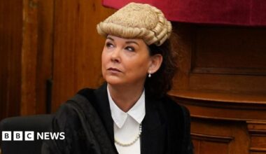 Dorothy Bain, who has long dark hair tied back under a court wig, looks up to her right while sitting down. She is wearing a black gown over a white shirt, with wood panelling behind her.