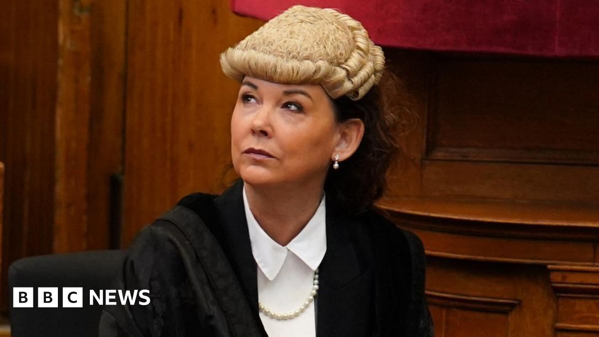 Dorothy Bain, who has long dark hair tied back under a court wig, looks up to her right while sitting down. She is wearing a black gown over a white shirt, with wood panelling behind her.