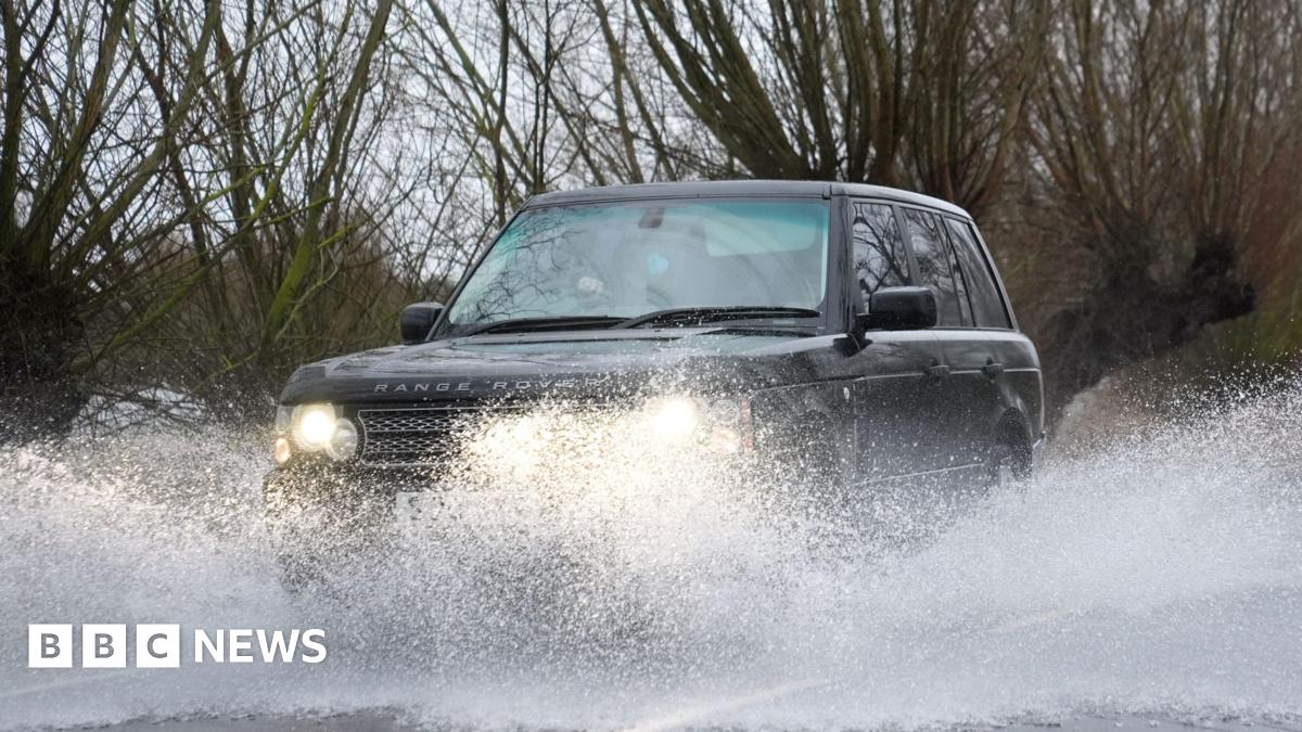 A large 4X4 car drives along a flooded road. Surface water is being catapulted into the air as the car drives through it. The Range Rover is a dark colour and has its headlights turned out. Trees are in the background.