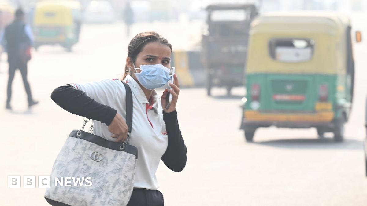 A person holds a phone to their ear as they walk down a street in Noida while wearing a face mask with tuk tuks in the background driving down the street, taken in November 2025.