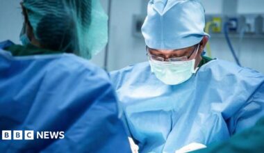 A surgeon wearing glasses and blue scrubs looks down at his hands as he operates on a patient