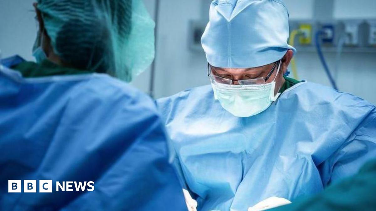 A surgeon wearing glasses and blue scrubs looks down at his hands as he operates on a patient