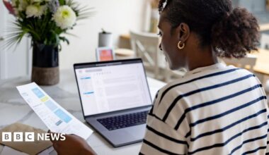 A woman looks at her bills in a letter and on a laptop whilst sat in her kitchen at home