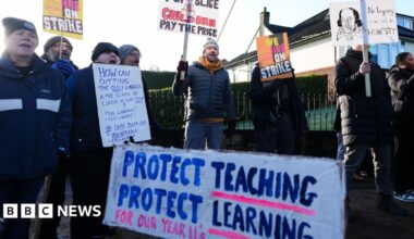 A group of people in winter clothing hold up sign supporting the strike action. A sign in the foreground reads: "Protect teaching, protect learning."