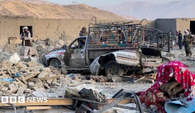 A heavily damaged vehicle with colourful lights installed on the sides is surrounded by rubble from a destroyed building. People stand around it inspecting the area. Mountains are in the distance.
