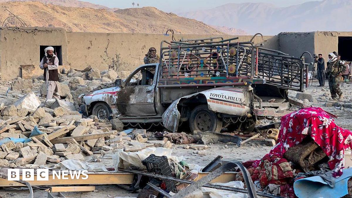 A heavily damaged vehicle with colourful lights installed on the sides is surrounded by rubble from a destroyed building. People stand around it inspecting the area. Mountains are in the distance.