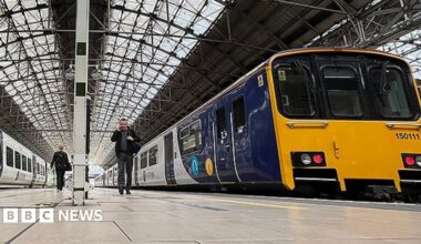 Platform level image of the inside of Manchester Piccadilly railway station with two yellow, blue and white Northern trains. A couple of passengers are walking to and from the trains.
