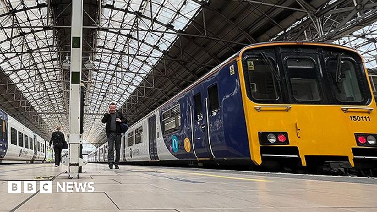 Platform level image of the inside of Manchester Piccadilly railway station with two yellow, blue and white Northern trains. A couple of passengers are walking to and from the trains.