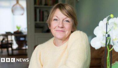 Annie sitting in a furnished room with a white flower in the foreground. She has short brown hair and is wearing a yellow jumper.