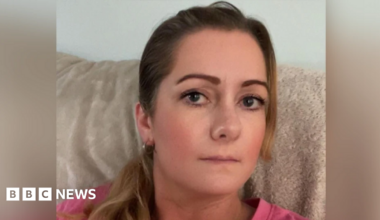 A white woman with dark hair and dark eyes lookiing at the camera, she is wearing a pink top